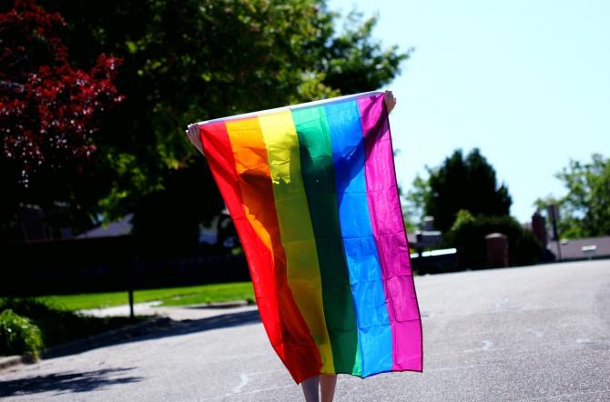 Person in silhouette holding up a sunlit rainbow Pride flag walking down a street