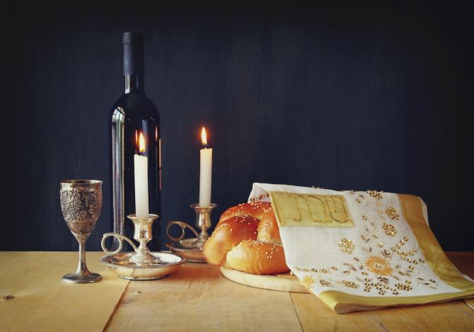 Table with wine bottle, kiddush cup, burning candles, covered challah