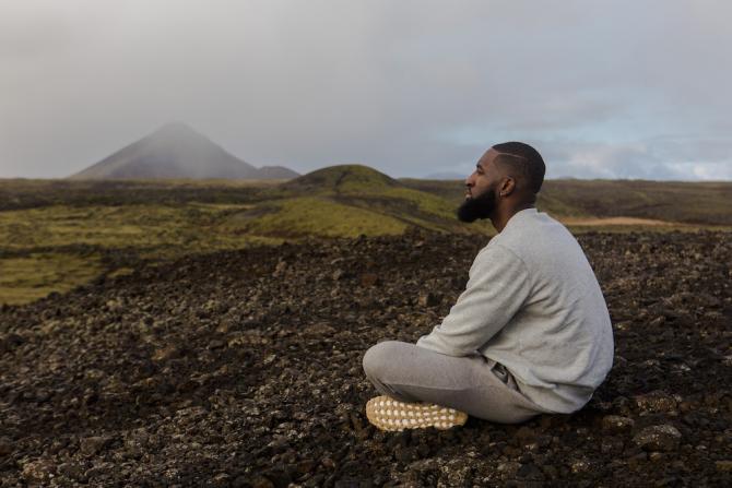 man sitting cross-legged outdoors meditating with mountain in background