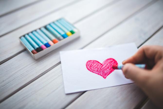 hand drawing a heart with pink crayon on a wooden table, with a box of multicolored crayons sitting nearby