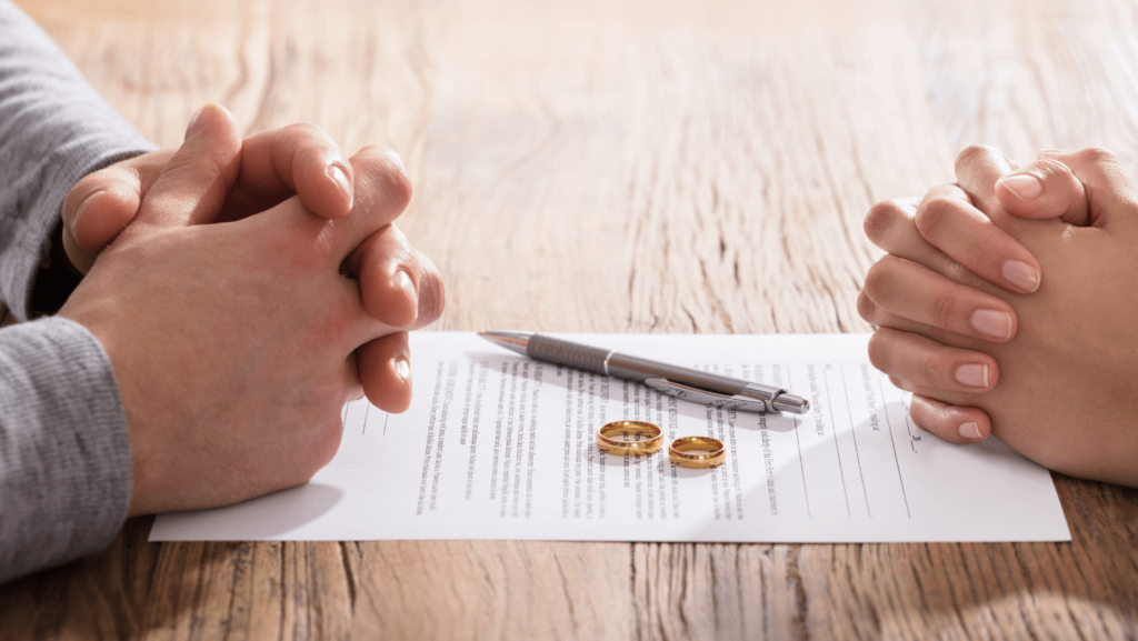 Two people sitting across from each other at a table, hands folded, with divorce papers and wedding rings between them