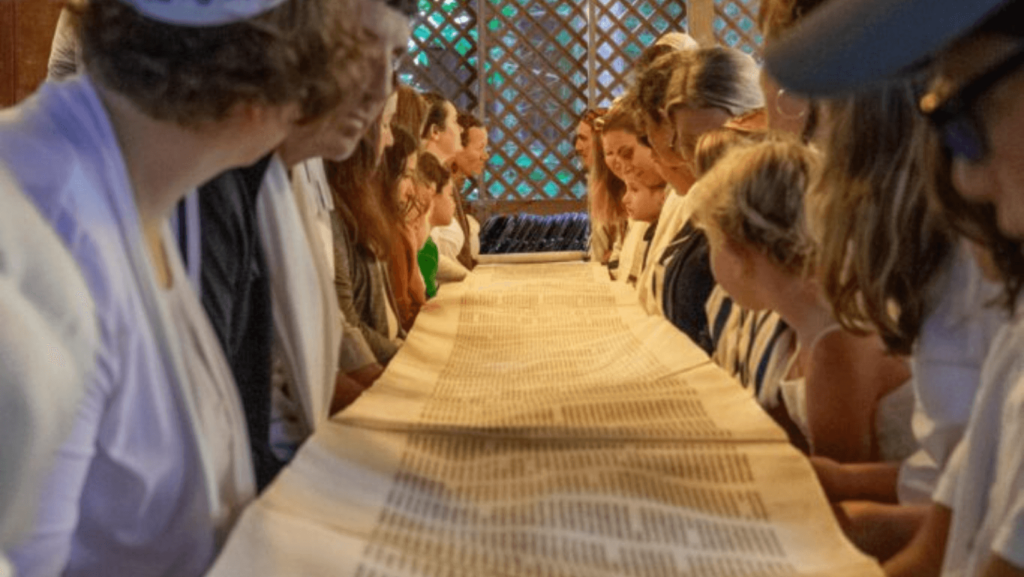 Multiple people gathered around either side of a long table, the Torah unrolled onto the table
